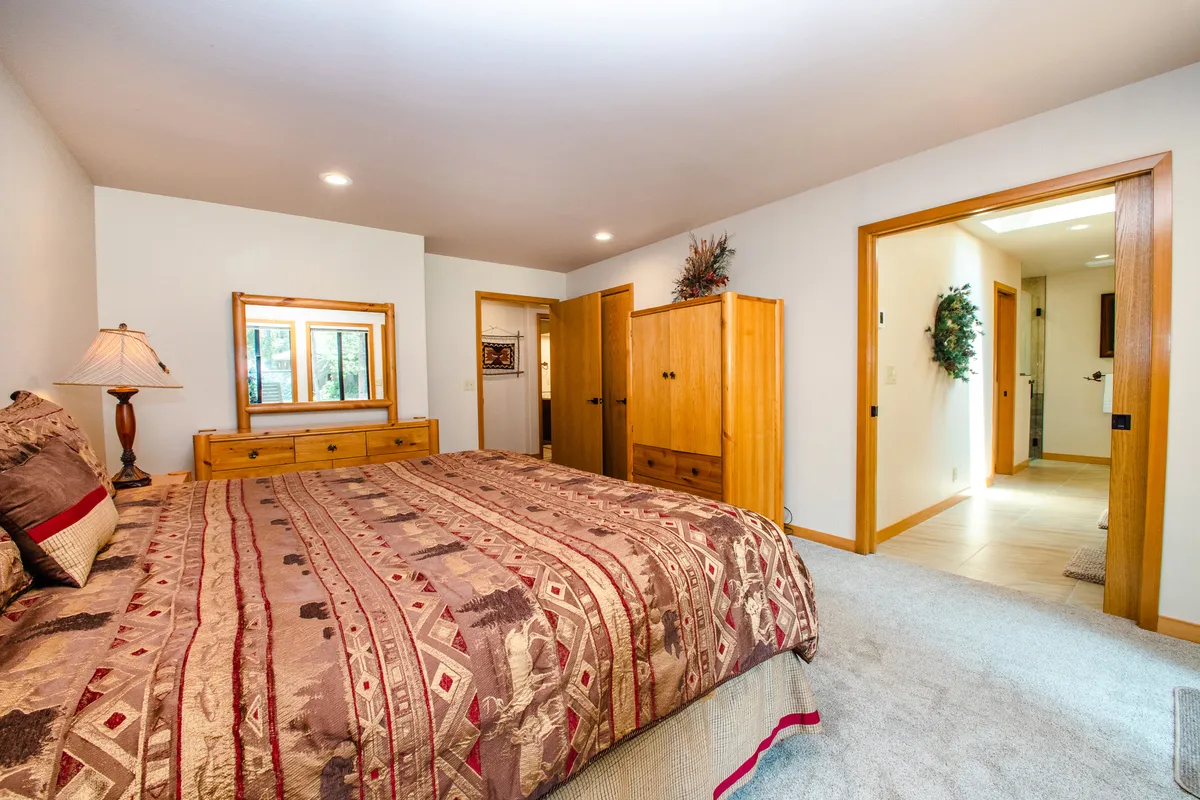 A cozy bedroom featuring a king-sized bed with patterned bedding, a wooden dresser with mirror, a bedside lamp, and wardrobe. Natural light streams in from large windows, creating a tranquil environment perfect for relaxation.