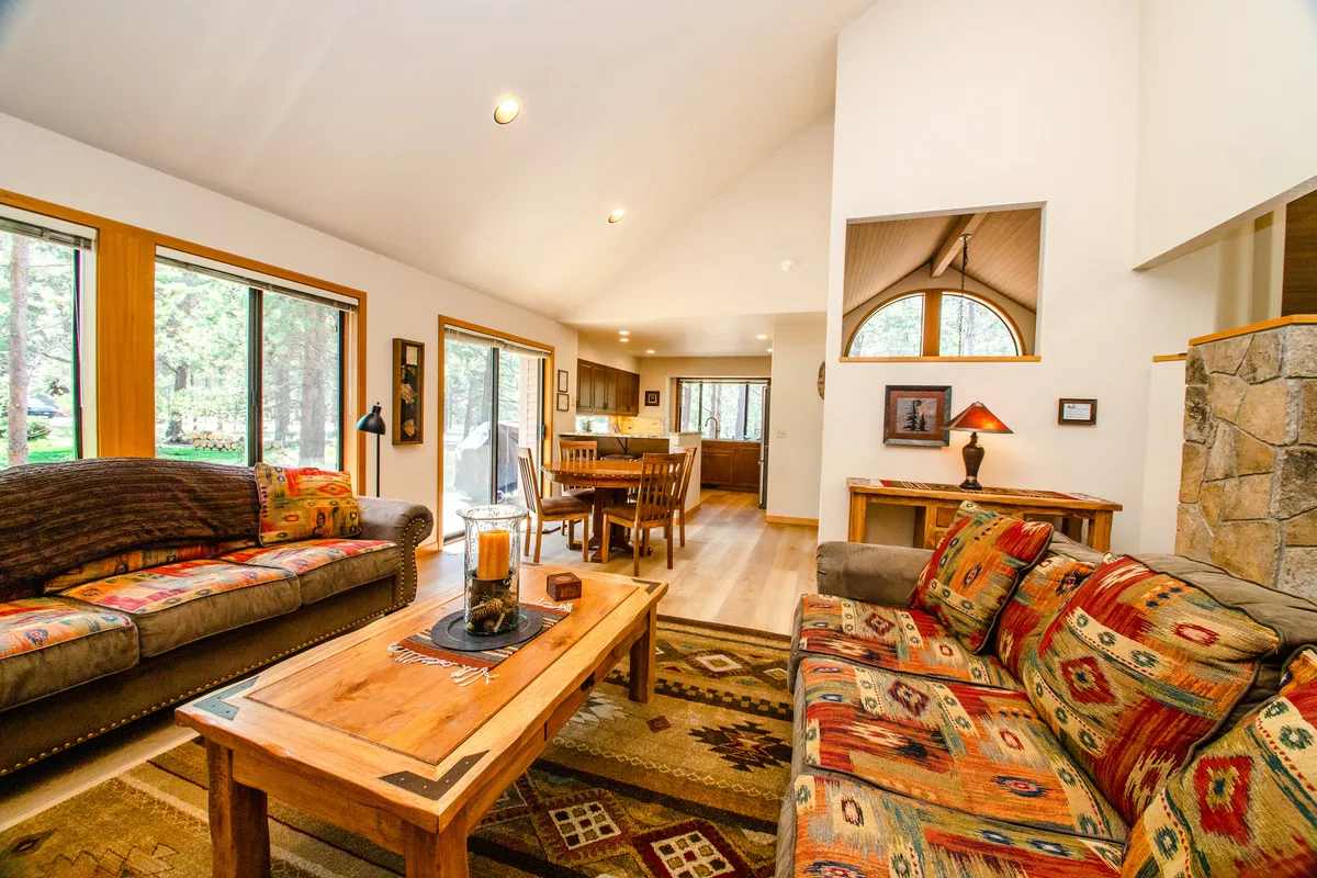 This image showcases a welcoming living area with colorful patterned sofas, a wooden coffee table, and large windows that let in natural light. The dining area is visible in the background, emphasizing the open-concept layout. The cozy furnishings and warm atmosphere invite relaxation and group gatherings.