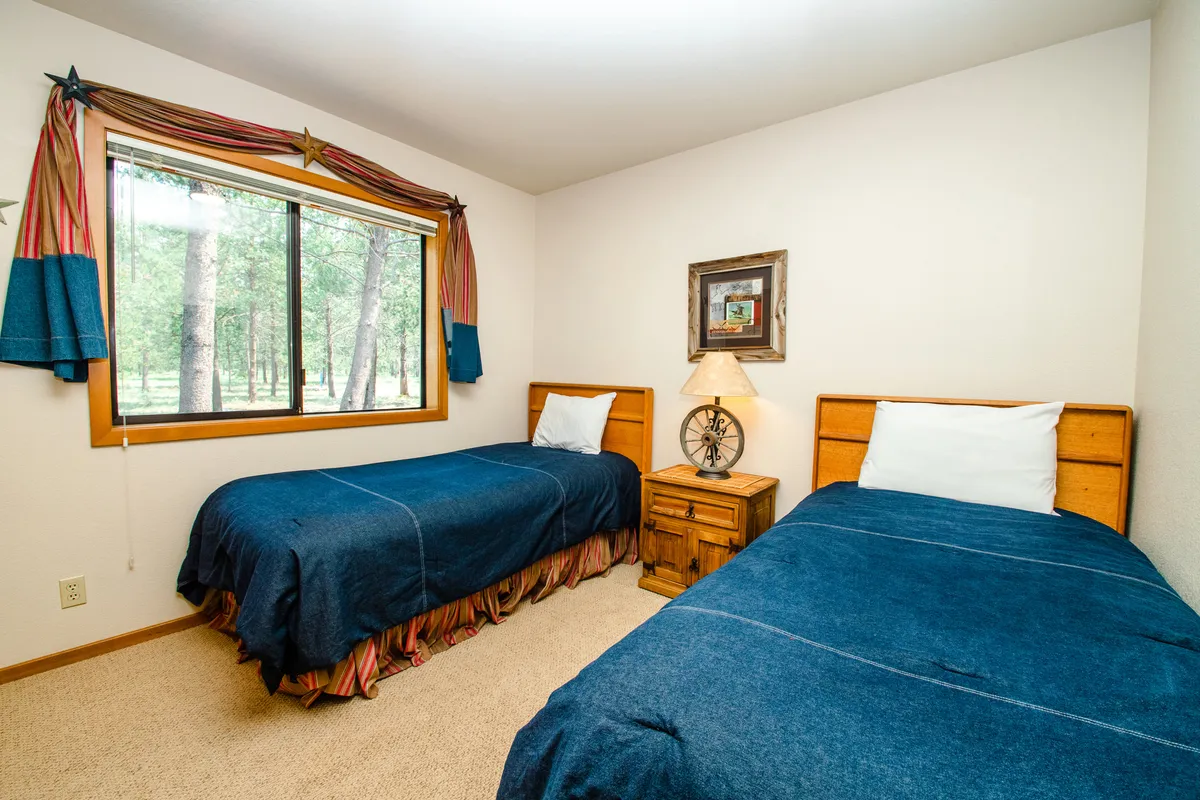 A comfortable bedroom with two twin beds, a nightstand with a lamp, framed artwork, and a window with a pleasant view. The soft lighting and neutral decor create a peaceful, versatile space suitable for children, friends, or couples.