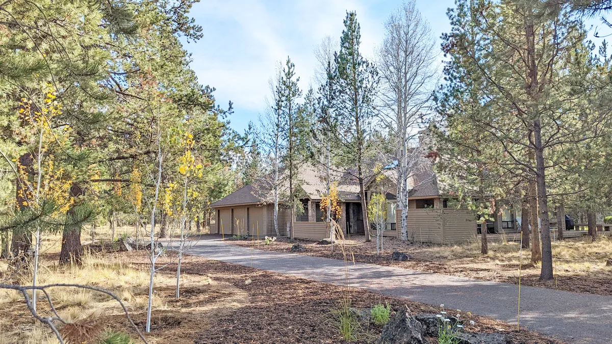 The image shows the home's main entrance framed by tall trees, a winding driveway, and welcoming landscaping. The natural surroundings create a serene and tranquil entry, ideal for arriving guests or groups gathering before an outing.