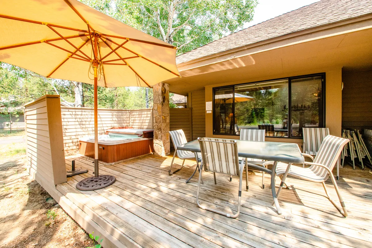 This exterior image captures a spacious deck complete with a bubbling hot tub and a dining table shaded by an umbrella. The deck is surrounded by greenery, creating a private, tranquil outdoor retreat ideal for relaxing, socializing, and enjoying meals in nature.