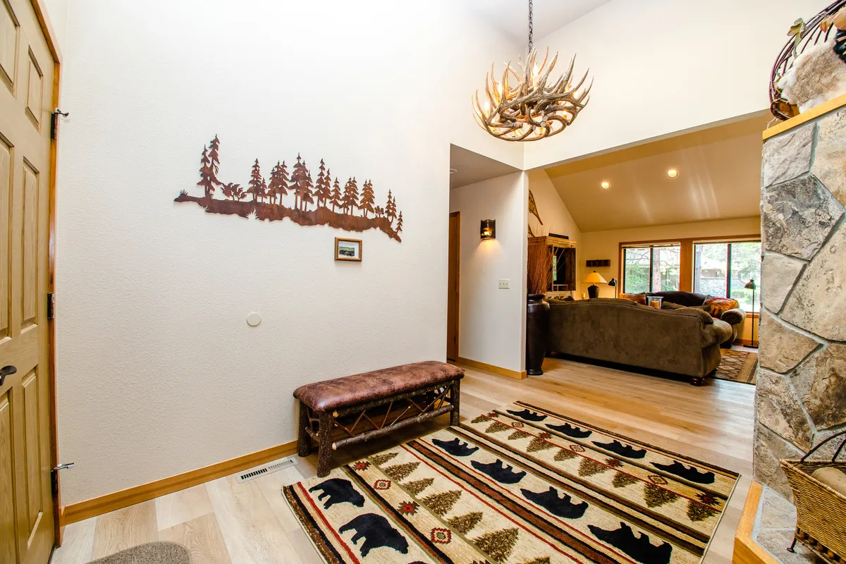 The entryway boasts a rustic antler chandelier, a wooden bench, a bear-themed rug, and wall art featuring trees, immediately evoking the sense of being in a mountain adventure basecamp. The atmosphere is cozy, welcoming, and full of outdoorsy character.