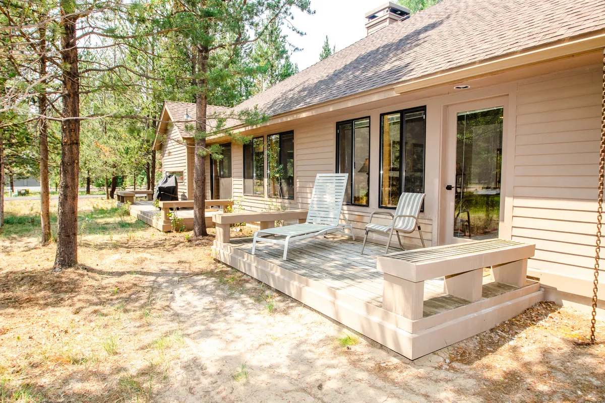 This exterior image features a spacious deck with comfortable seating overlooking lush trees and greenery. Large windows reflect natural light, and the setting is tranquil, inviting, and perfect for outdoor relaxation or group gatherings.