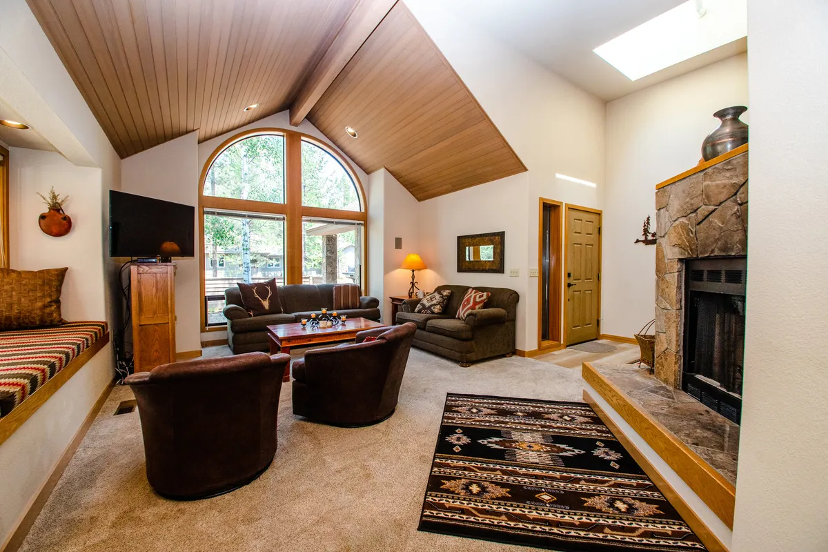 The image showcases a bright, inviting living room featuring large arched windows that bathe the space in natural light, a warm fireplace as the focal point, and plush, textured seating arranged around a decorative rug—perfect for gathering and relaxing.